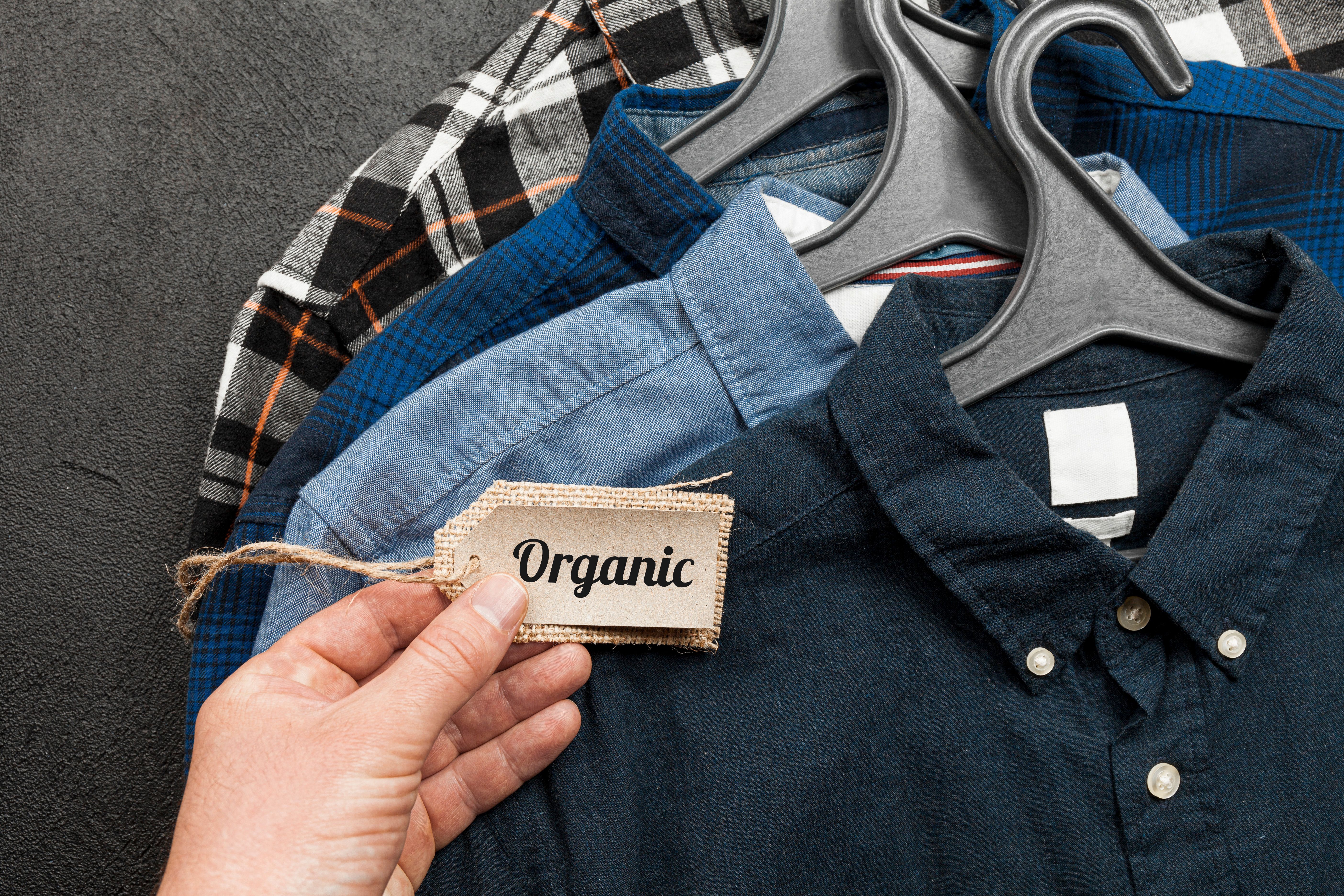 Hand holding organic label in front of eco-friendly cotton shirts on hangers – sustainable and ethical clothing choice
