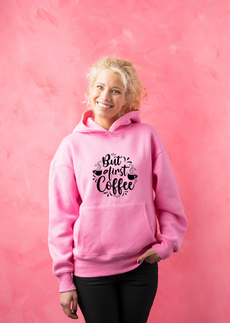 Smiling woman in a pink hoodie with 'But first coffee' text and coffee motif, standing in front of a pink wall – customize your hoodie