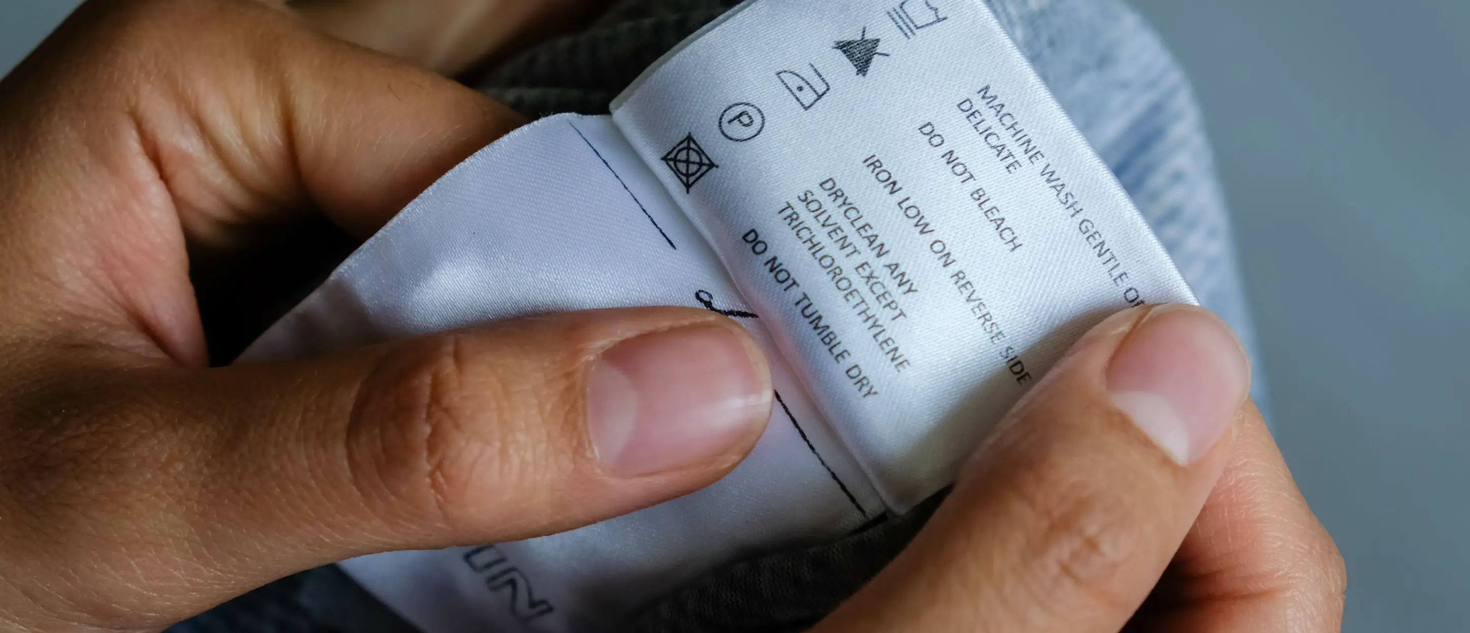 Hände halten ein Kleidungsetikett mit Pflegehinweisen wie Waschen, Bügeln und Reinigung – Nahaufnahme Textiletikett