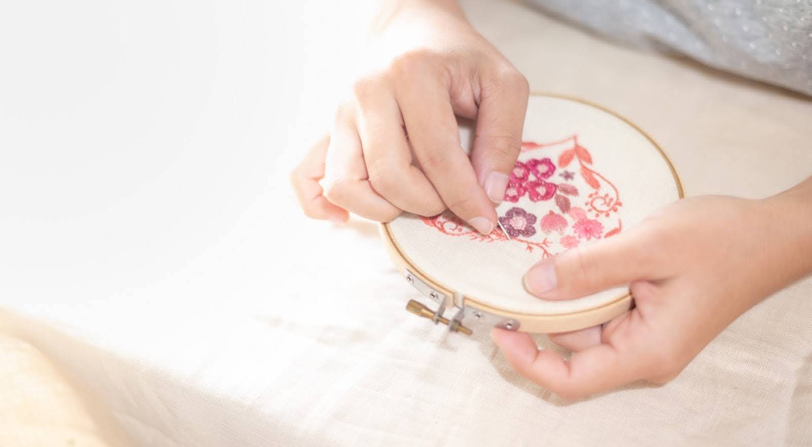 Frau stickt ein rosa Blumenmuster mit Nadel und Stickrahmen auf Stoff – filigrane Handarbeit in der Entstehung
