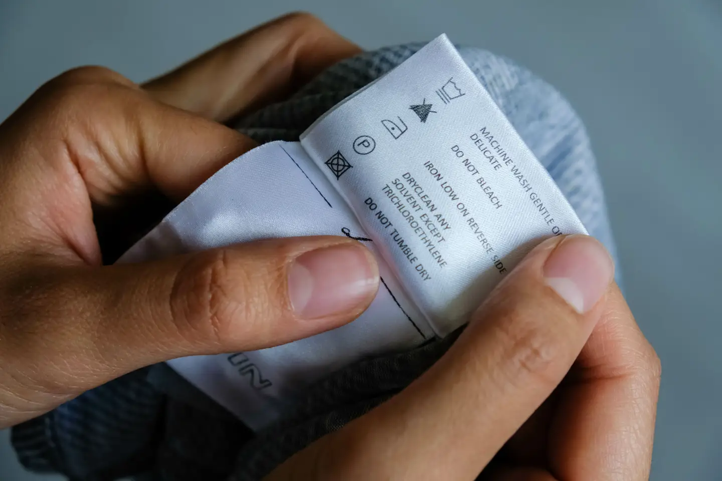 Hände halten ein Kleidungsetikett mit Pflegehinweisen wie Waschen, Bügeln und Reinigung – Nahaufnahme Textiletikett