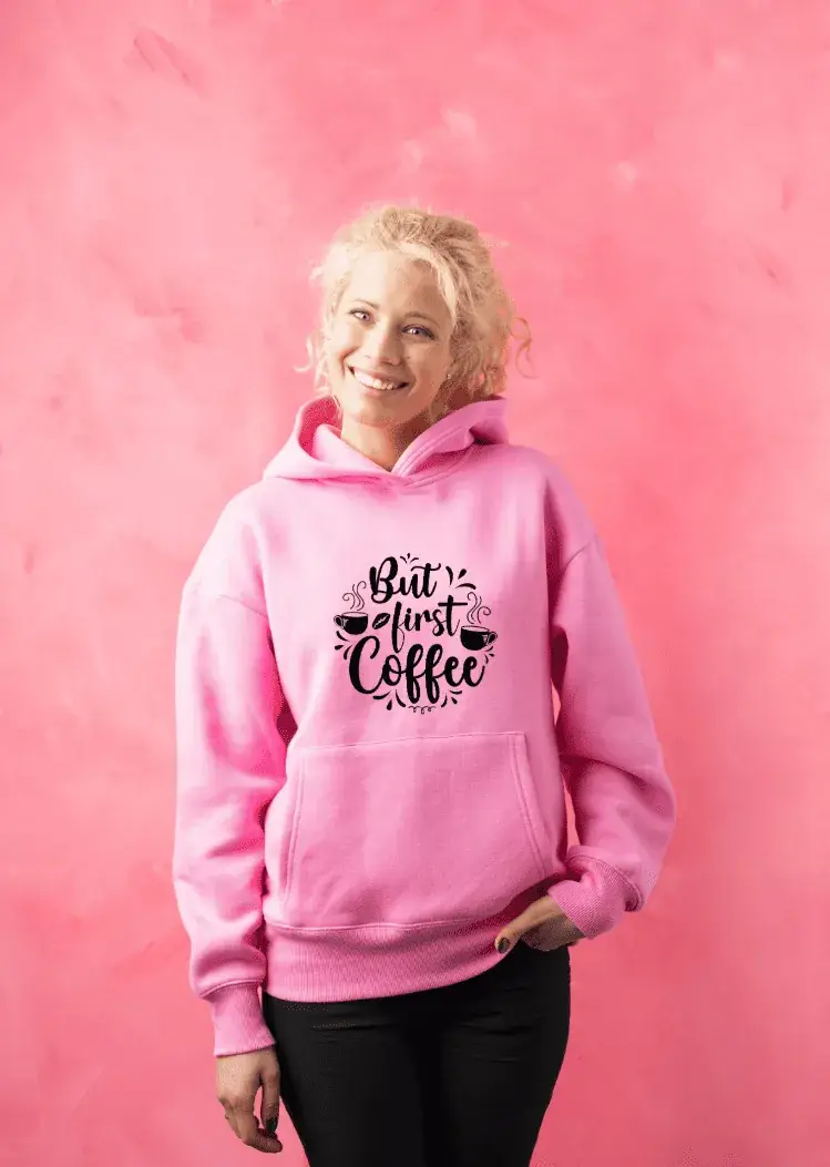 Blonde Frau im pinken Hoodie mit 'But first coffee'-Design Lachende Frau trägt pinken Hoodie mit 'But first coffee'-Spruch und Kaffeemotiv, steht vor pinker Wand – Hoodie individuell gestalten