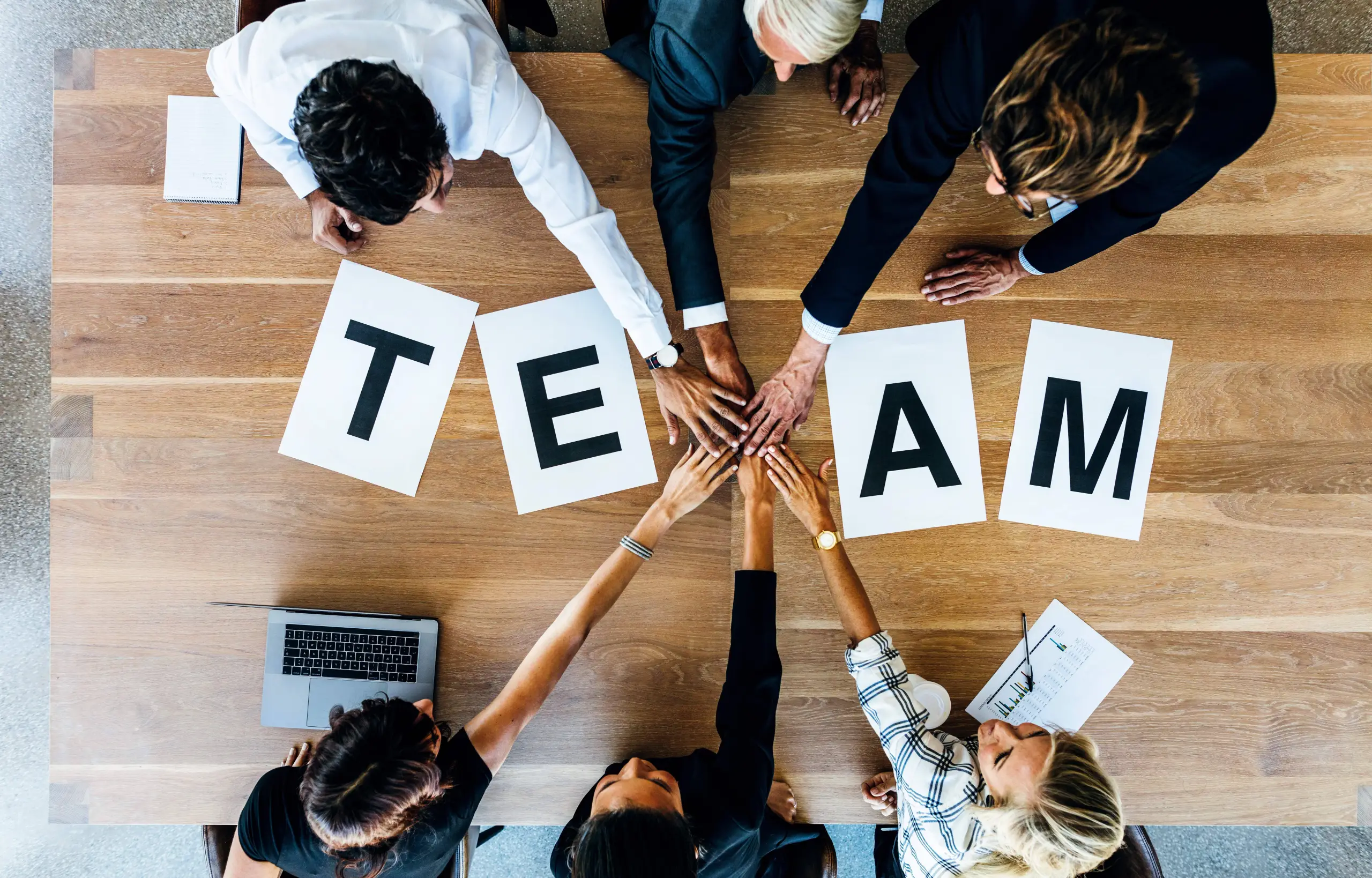 Teamwork mit Firmenbekleidung – Hände gestapelt über dem Wort „Team“, T-Shirts für Teambuilding.